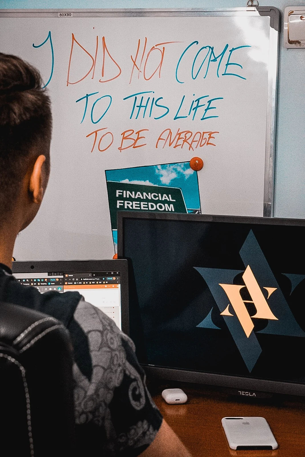 Andrei at his desk, photographed from behind over his shoulder. On the whiteboard in front of him, handwritten in blue and orange capitals, is the line I DID NOT COME TO THIS LIFE TO BE AVERAGE. A postcard of a green road sign reading FINANCIAL FREEDOM is pinned underneath it. An open laptop and a monitor sit on the desk.