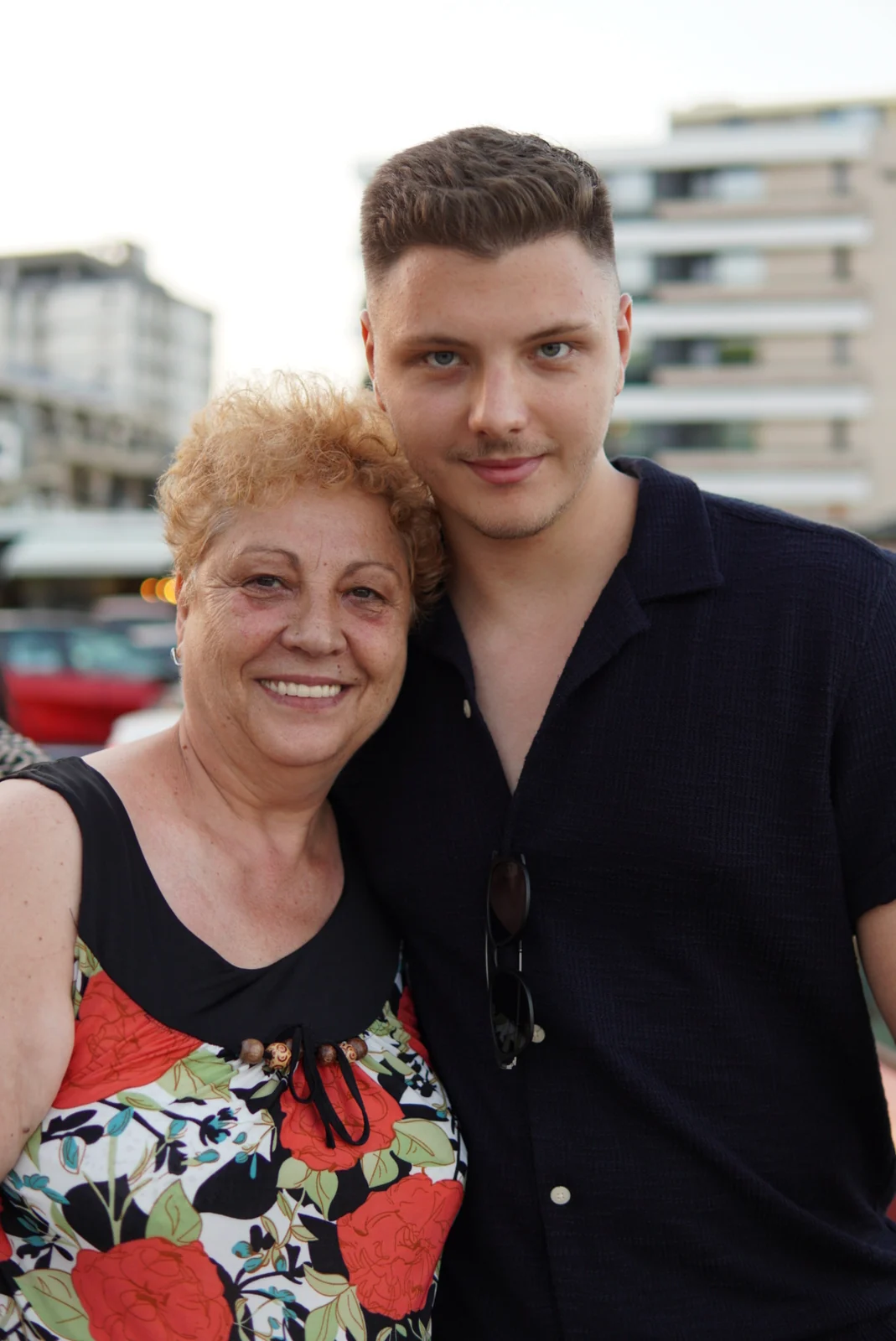 Andrei standing cheek to cheek with his mother outdoors, both smiling at the camera. She has short fair hair and a floral dress. He is in a dark shirt with sunglasses hanging from the collar.